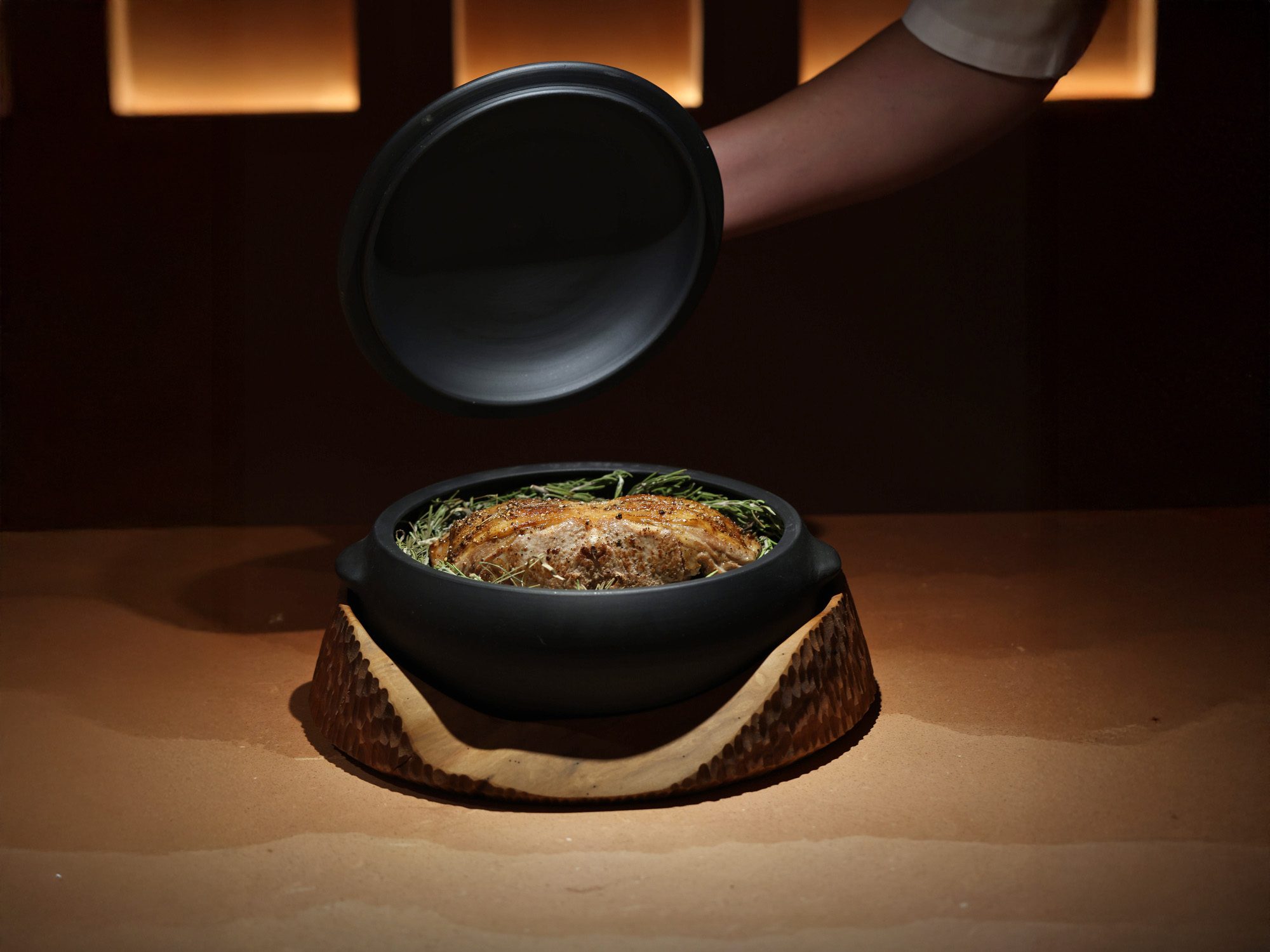Panela de barro preto sobre suporte de madeira entalhada, com carne assada e ervas, a tampa erguida à mesa