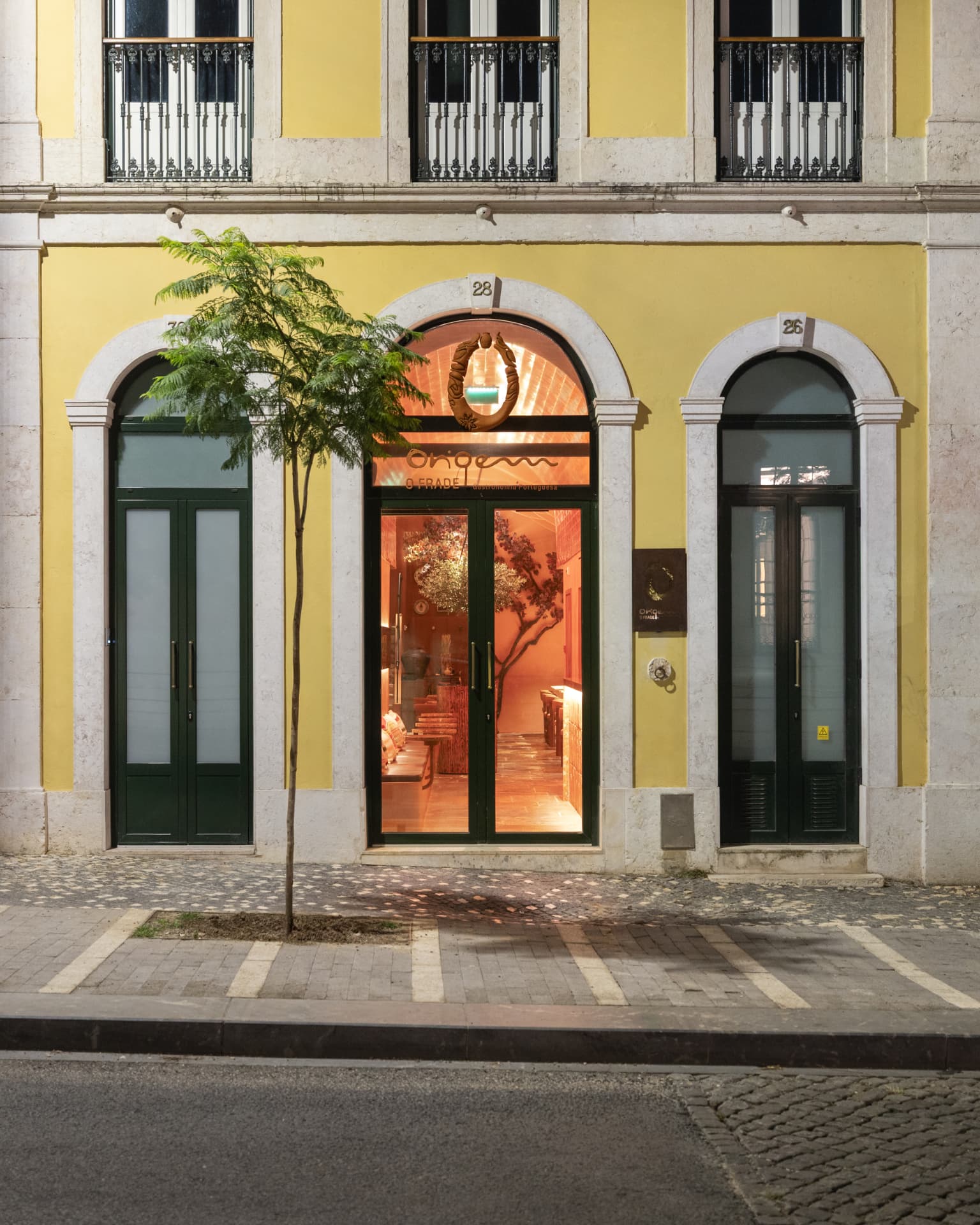 Fachada do Origem à noite, com portas verdes, letreiro dourado e luz quente junto à Calçada da Ajuda