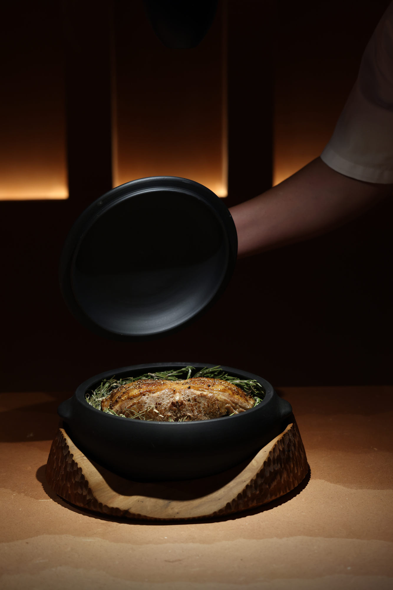 Clay lid lifted over roast meat on a bed of herbs